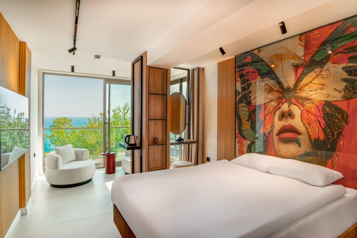A modern bedroom with a large bed, a seating area with a chair and a table, a large window with a view of the ocean, and a colorful abstract painting on the wall.