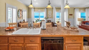 A kitchen with a large island, wooden cabinets, and a dishwasher. There are two pendant lights, a view of the ocean through large windows, and a dining area with a table and chairs. - Perfect Timing- On The Beach, Sleeps 10 (Port St. Joe)