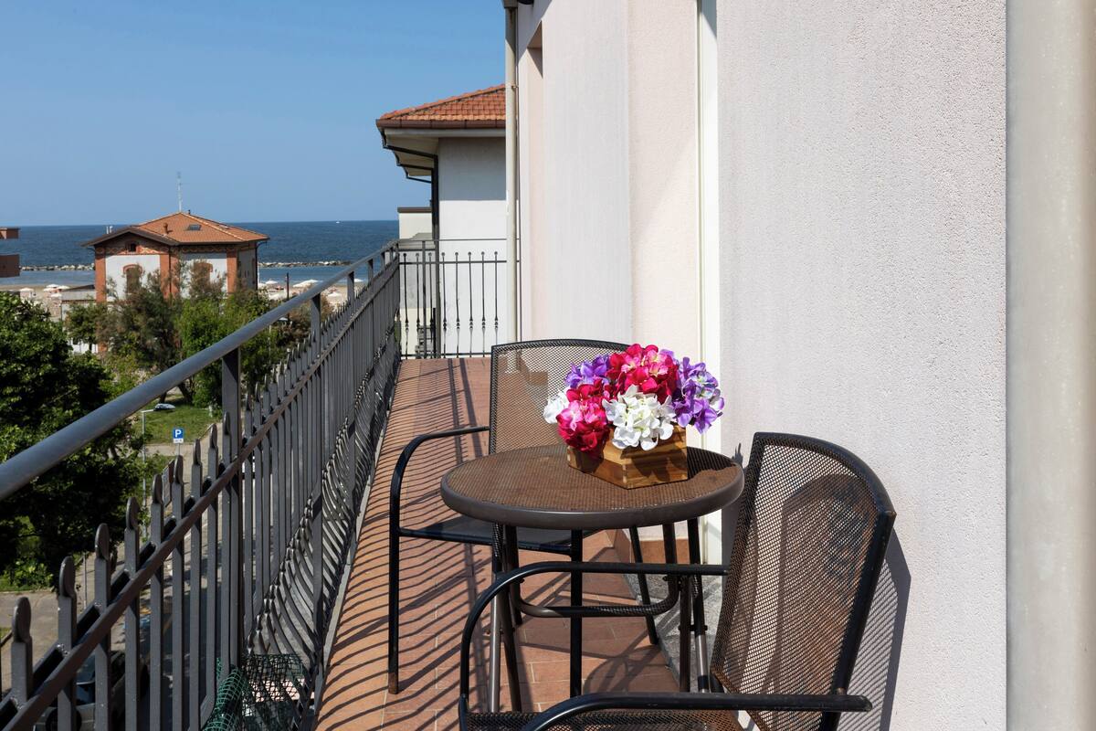 Un balcon avec une table et des chaises, un vase de fleurs et une vue sur la mer et les bâtiments.