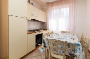 A compact kitchen with a gas stove, oven, and sink, a dining table with chairs, and a window with sheer curtains. - Residence 3 Trilo&Suites (Rimini)