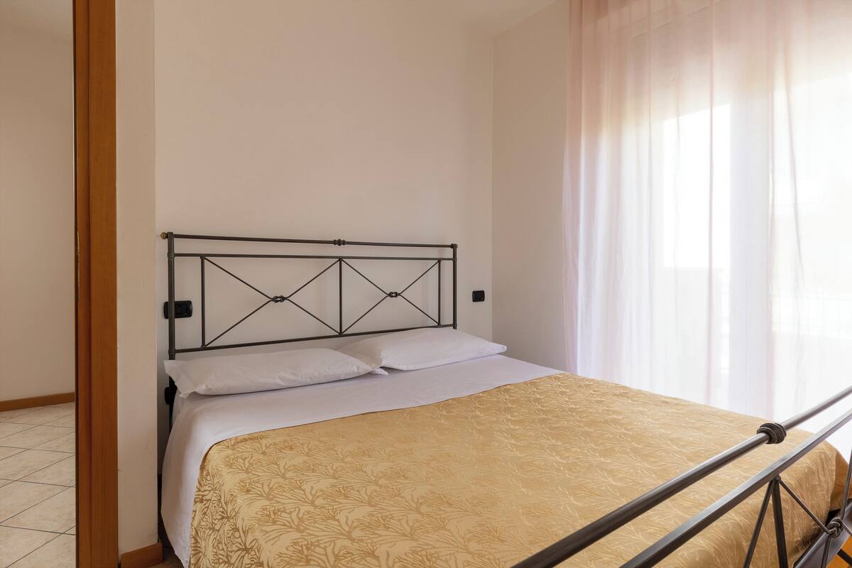 Un lit avec un cadre en métal et une couvre-lit à motifs, dans une pièce aux murs unis et dotée d’une fenêtre ornée de rideaux légers.