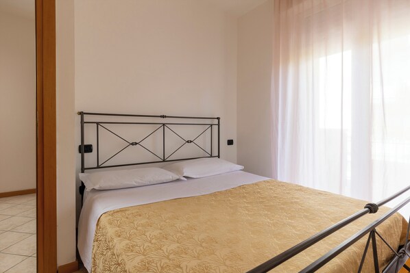 A bed with a metal frame and a patterned bedspread in a room with plain walls and a window with sheer curtains. - Residence 3 Trilo&Suites (Rimini)