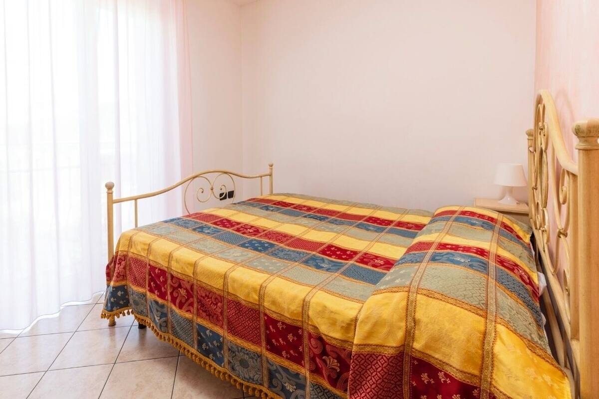 Un lit simple avec une courtepointe, une tête de lit en bois, une table de chevet avec une lampe et une fenêtre avec des rideaux légers.