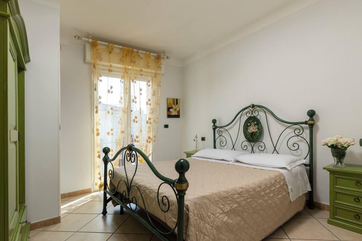 Une pièce avec un lit, une table de chevet verte, une fenêtre avec des rideaux et un tableau accroché au mur.