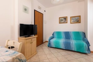 A room with a television on a wooden cabinet, a blue-striped sofa, a wooden door, and two framed pictures on the wall. - Residence 3 Trilo&Suites (Rimini)