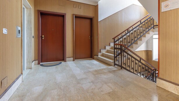 Un vestíbulo de hotel con piso de mármol, paneles de madera, dos puertas de madera y una escalera con barandilla metálica.