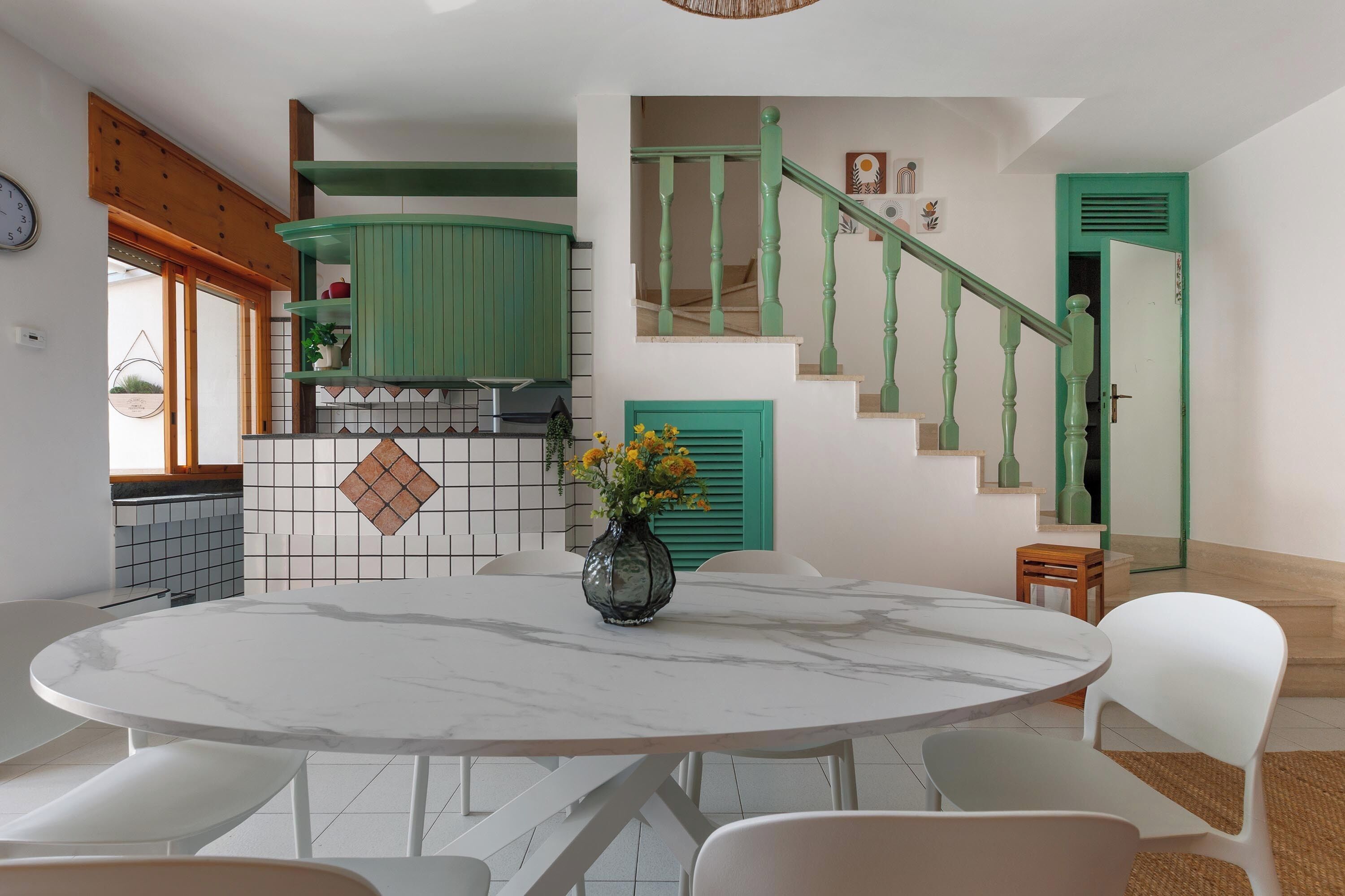 A dining area with a marble table and white chairs, a kitchen with green accents, and a staircase with green railings.