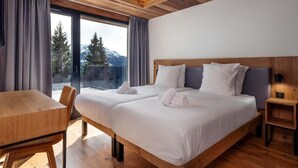 A modern hotel room with a large bed, a wooden table, and a balcony with a view of trees and mountains.