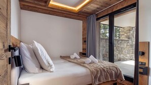 A bedroom with a wooden ceiling, a bed with white linens, and a view of a stone wall through a glass door.