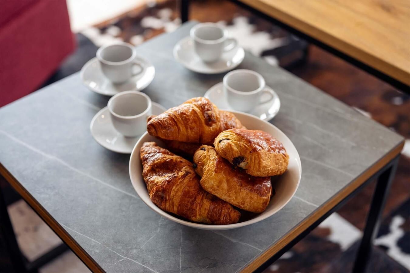 Una mesa con un bol de cruasanes y cuatro tazas blancas sobre platillos.