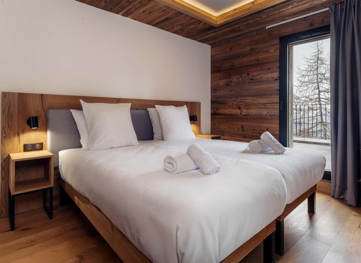 A modern bedroom with a wooden headboard, a large bed, a nightstand, and a window with a view of trees.