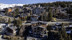 Una zona turística montañosa con varios edificios de varios pisos, probablemente hoteles, rodeada de picos nevados y un bosque denso.