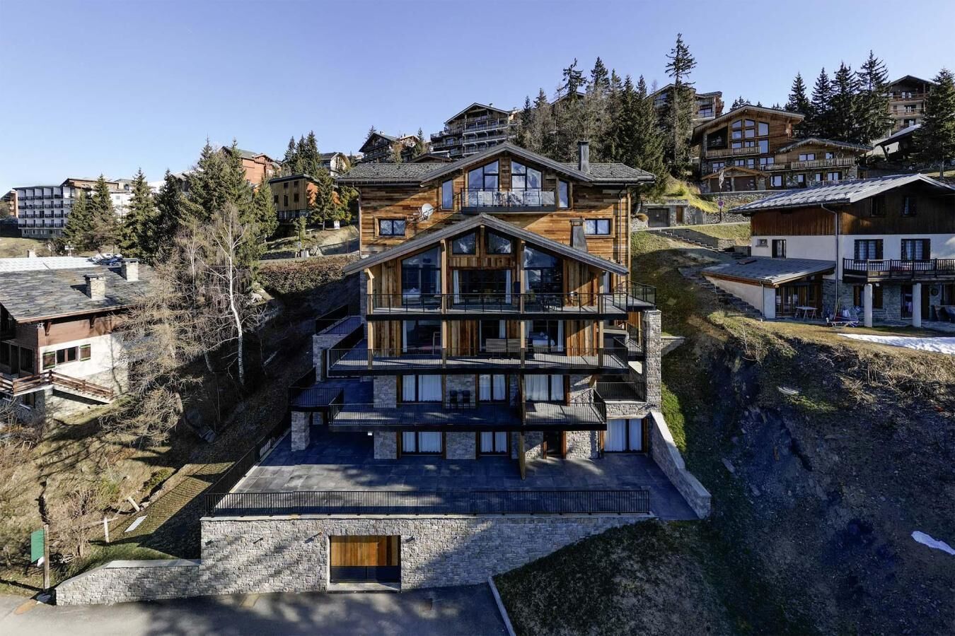 A multi-story wooden chalet with balconies, set in a mountainous area with other buildings in the background.