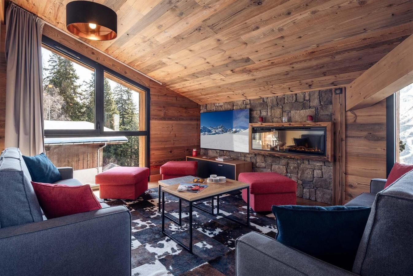 Un accogliente interno di una baita con camino in pietra, soffitto in legno e ampie finestre che si affacciano su alberi.