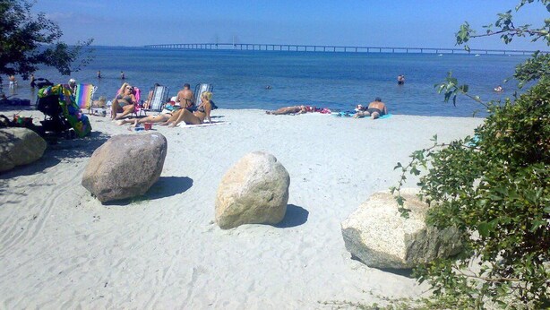 A beach with large rocks, clear blue water, and a bridge in the distance.
