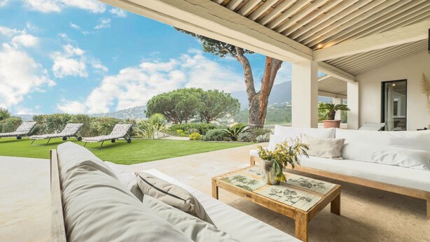 A covered outdoor seating area with a sofa and a coffee table, a lawn with sun loungers, and a view of trees and mountains. - Villa Maeva in Cavalaire-sur-mer (Cavalaire-sur-Mer)