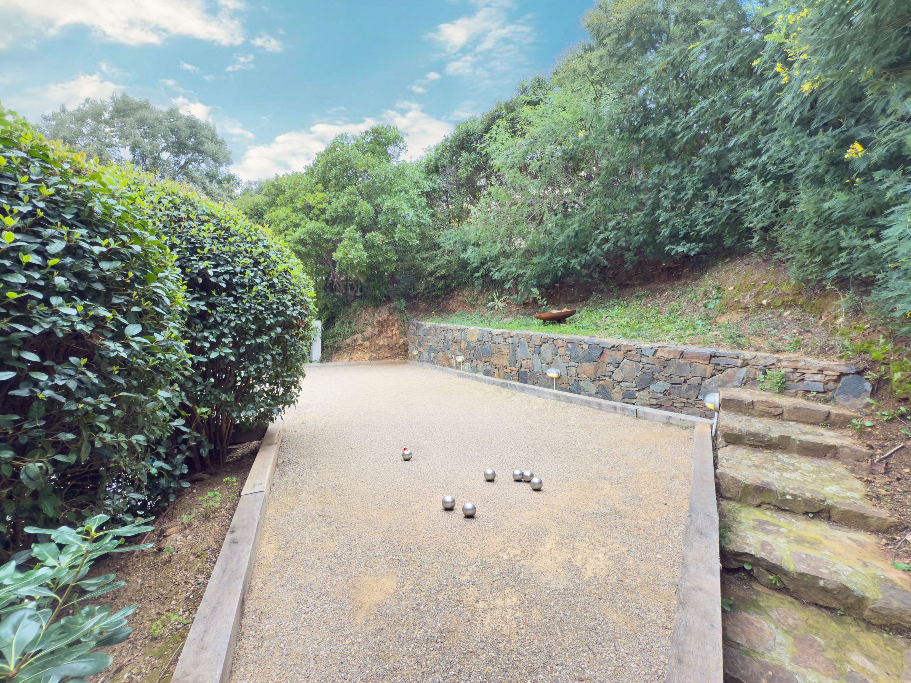 Un terrain de croquet recouvert de gravier, entouré d’une végétation luxuriante et bordé d’un mur de pierre.