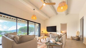 A spacious living room with a large window, a ceiling fan, and a flat-screen TV. - Villa Maeva in Cavalaire-sur-mer (Cavalaire-sur-Mer)