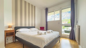 A neatly made bed with white linens and rolled towels, a bedside table with a lamp, a red chair, and a view of the outdoors through a glass door. - 213 Le Montfleury in Cannes (Cannes)
