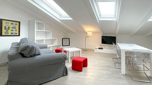 A modern loft apartment with a high ceiling, skylights, a grey sofa, a red ottoman, a white dining table, and a TV mounted on the wall. - 210 - Tony Allard 4A (Cannes)