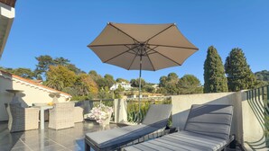 Ein sonniger Außenbereich mit Sitzgelegenheit, einer Liege, einem Tisch, einer Topfpflanze und einem großen Sonnenschirm.