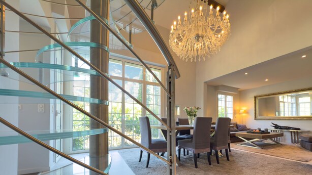 Ein modernes Interieur mit einer gläsernen Wendeltreppe, einem Essbereich mit Stühlen und Tisch sowie einem Kronleuchter.