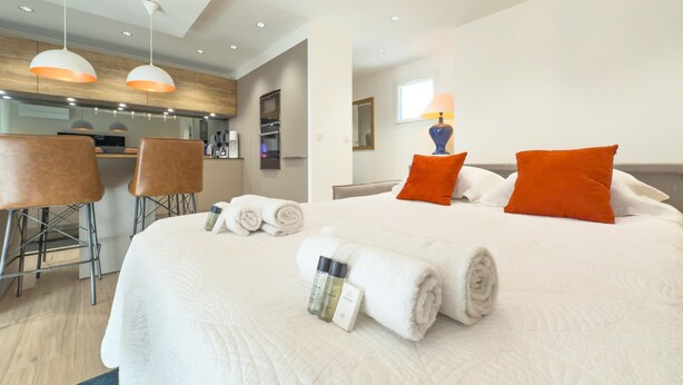 A modern hotel room with a large bed, a sitting area with a bar, and a kitchenette.
