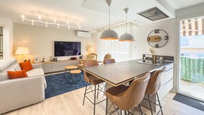 A modern living room with a sofa, a dining area with a table and chairs, a TV mounted on the wall, pendant lights, and a large window.