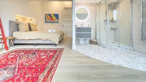 A modern bedroom with a bed, a red patterned rug, a shower with a glass door, and a bathroom with a vanity and mirror.