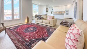 A spacious living room with a large window, a beige sofa, a patterned rug, and a kitchen in the background.