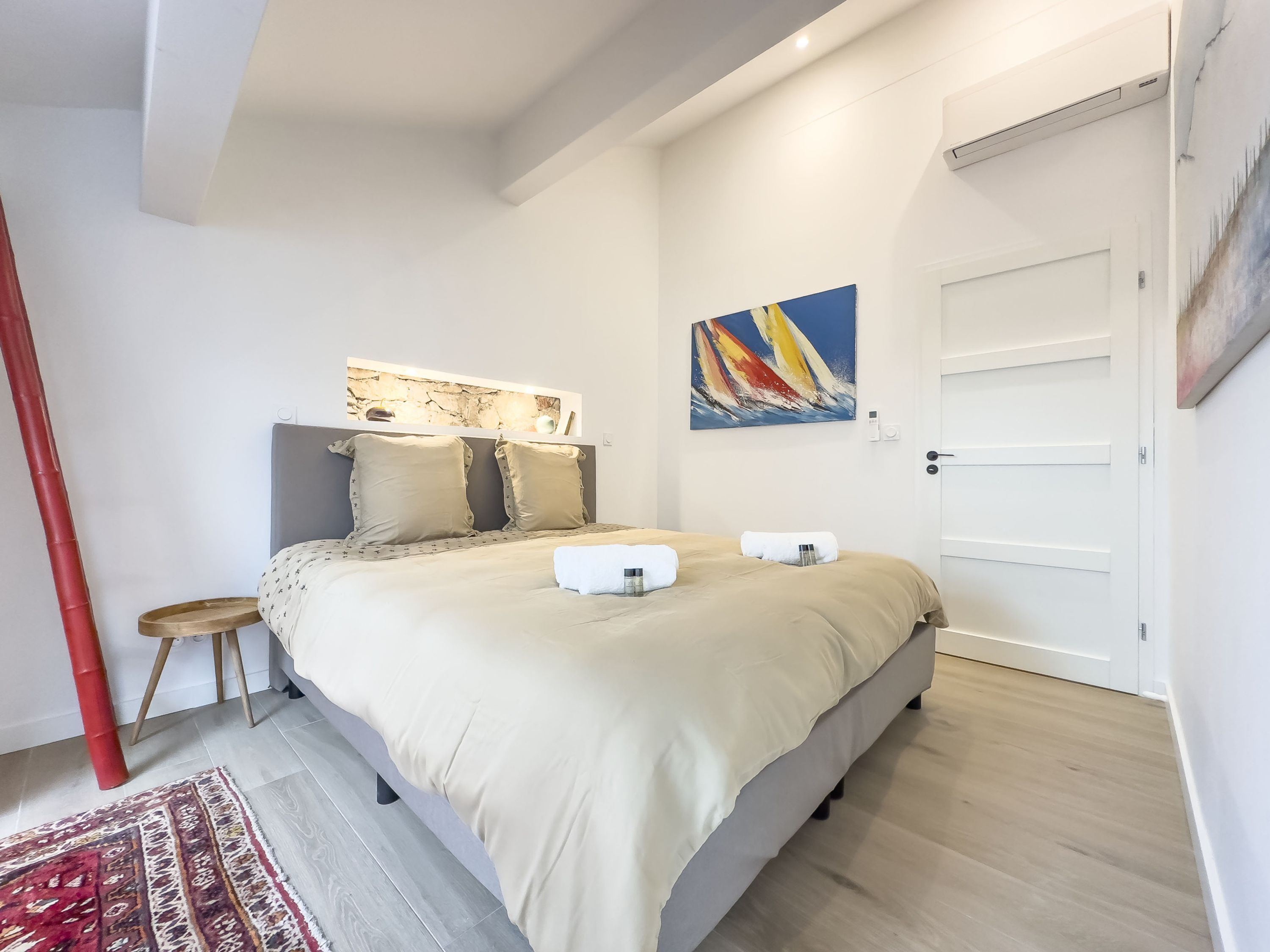 A modern bedroom with a large bed, a small stool, a red pole, a patterned rug, and a painting on the wall.