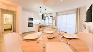 Una cocina moderna con electrodomésticos integrados, una zona de comedor con una mesa redonda para cuatro personas y un salón con sofá y televisor.