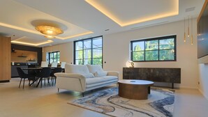 A modern living room with a sofa, coffee table, and a rug. There is a dining area with chairs and a table. Large windows provide natural light and views of greenery. - 007 Villa Deva in Cannes (Cannes)