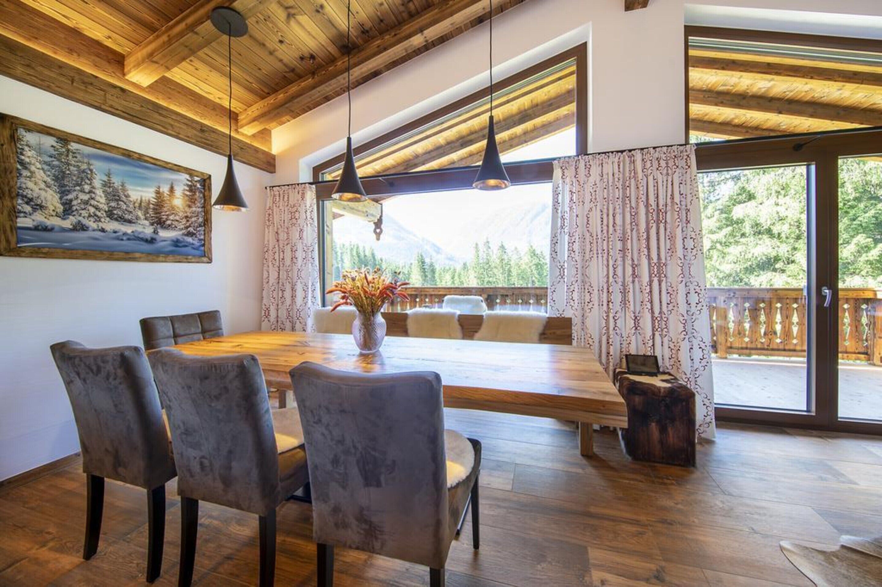 A dining area with a wooden table and chairs, a vase with flowers, and a view of a balcony with a wooden railing.