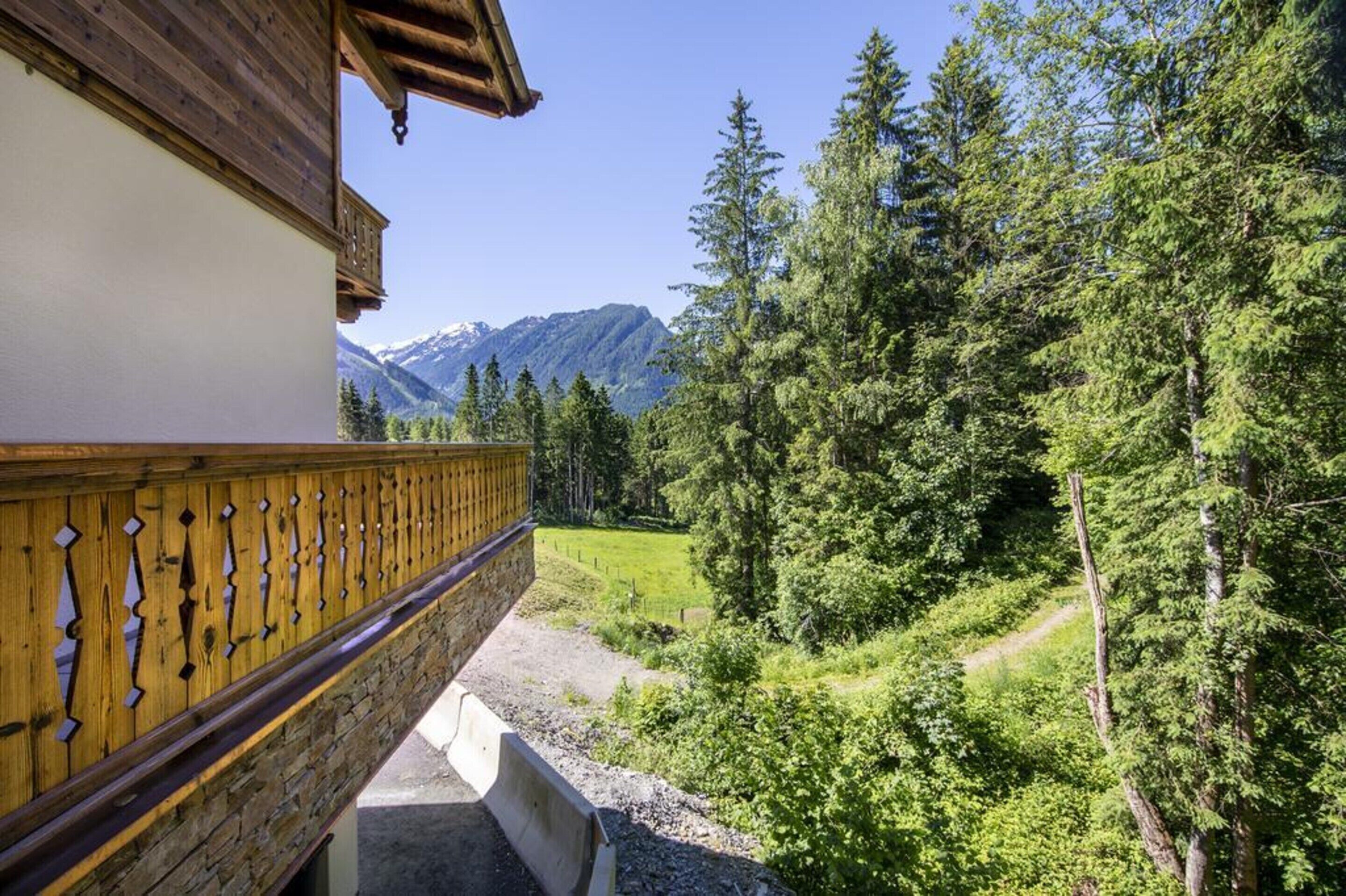 A balcony with a stone wall and wooden railing overlooking a forested valley with mountains in the background.