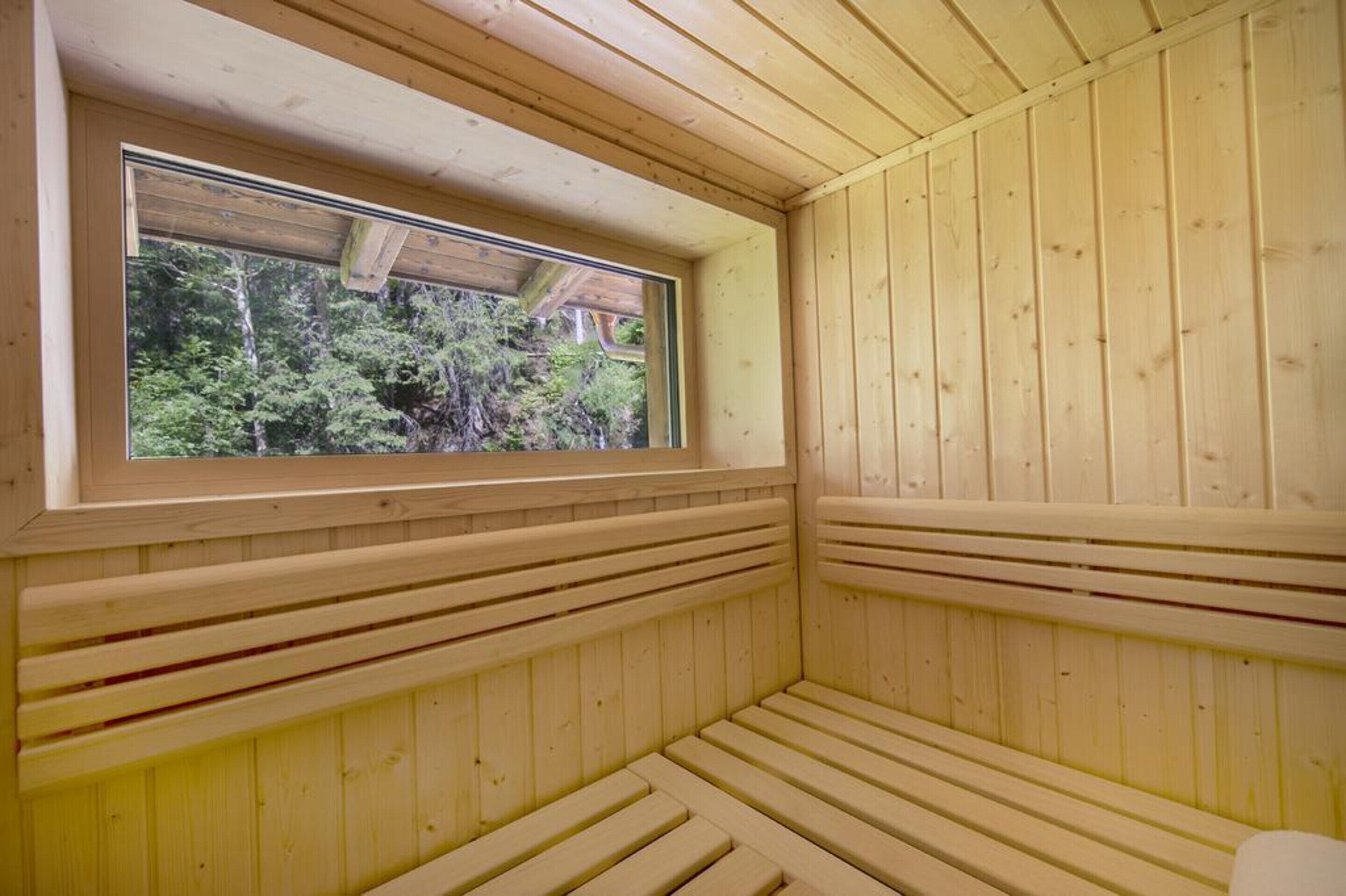 Un cuarto de sauna con paredes y bancos de madera, un ventanal con vistas a árboles y un techo de madera.