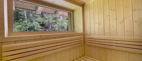 Un cuarto de sauna con paredes y bancos de madera, un ventanal con vistas a árboles y un techo de madera.