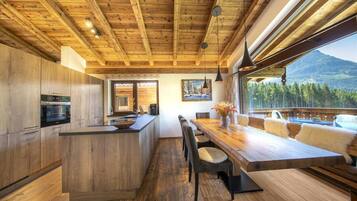 A modern kitchen with wooden cabinets and a central island, a dining area with a wooden table and chairs, and a view of a forested mountain range through large windows.