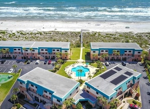 Um hotel à beira-mar com vários edifícios, uma piscina central e vista para a praia.