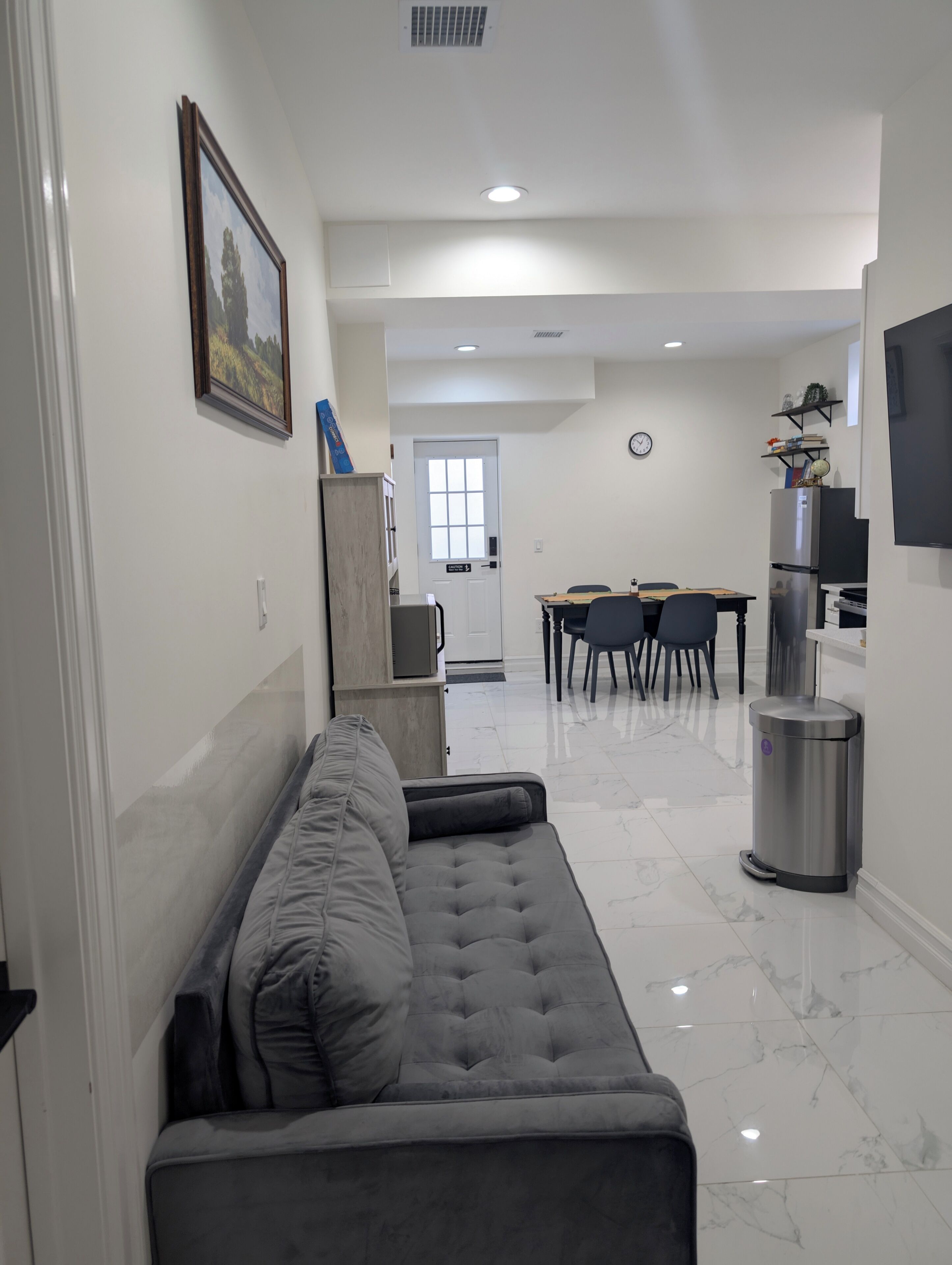 Una sala de estar moderna con un sofá gris, un comedor con mesa y sillas, un área de cocina con refrigerador, y un televisor montado en la pared.