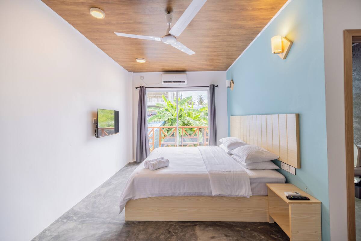 Une chambre moderne avec un lit en bois, un ventilateur de plafond, une table de chevet et une vue sur un balcon agrémenté de plantes.