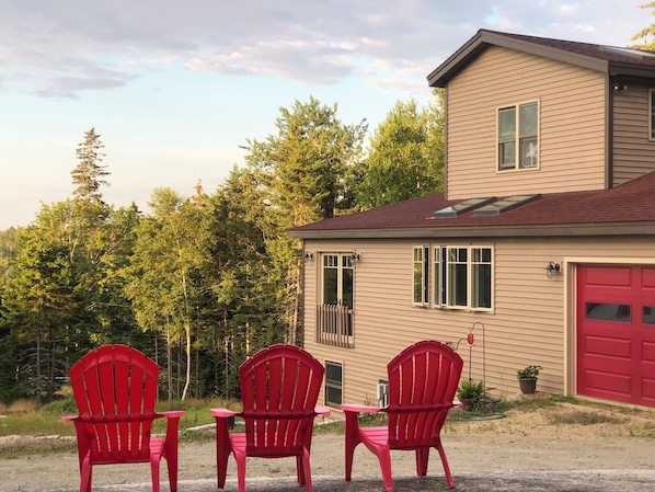 A house with a red garage door, a small attached garage, and a covered porch area with three red Adirondack chairs. - Oceanville Oasis on the Peaceful Island (Stonington)