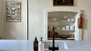 Une cuisine avec une cheminée en briques blanches, une estampe botanique encadrée et un panneau au-dessus de la cheminée indiquant différents sports.