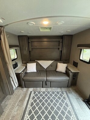 A compact RV interior with a sofa bed, two side tables, a window, and a patterned rug. - Luxury Meets Adventure. Glamp in Style. (Roscommon)