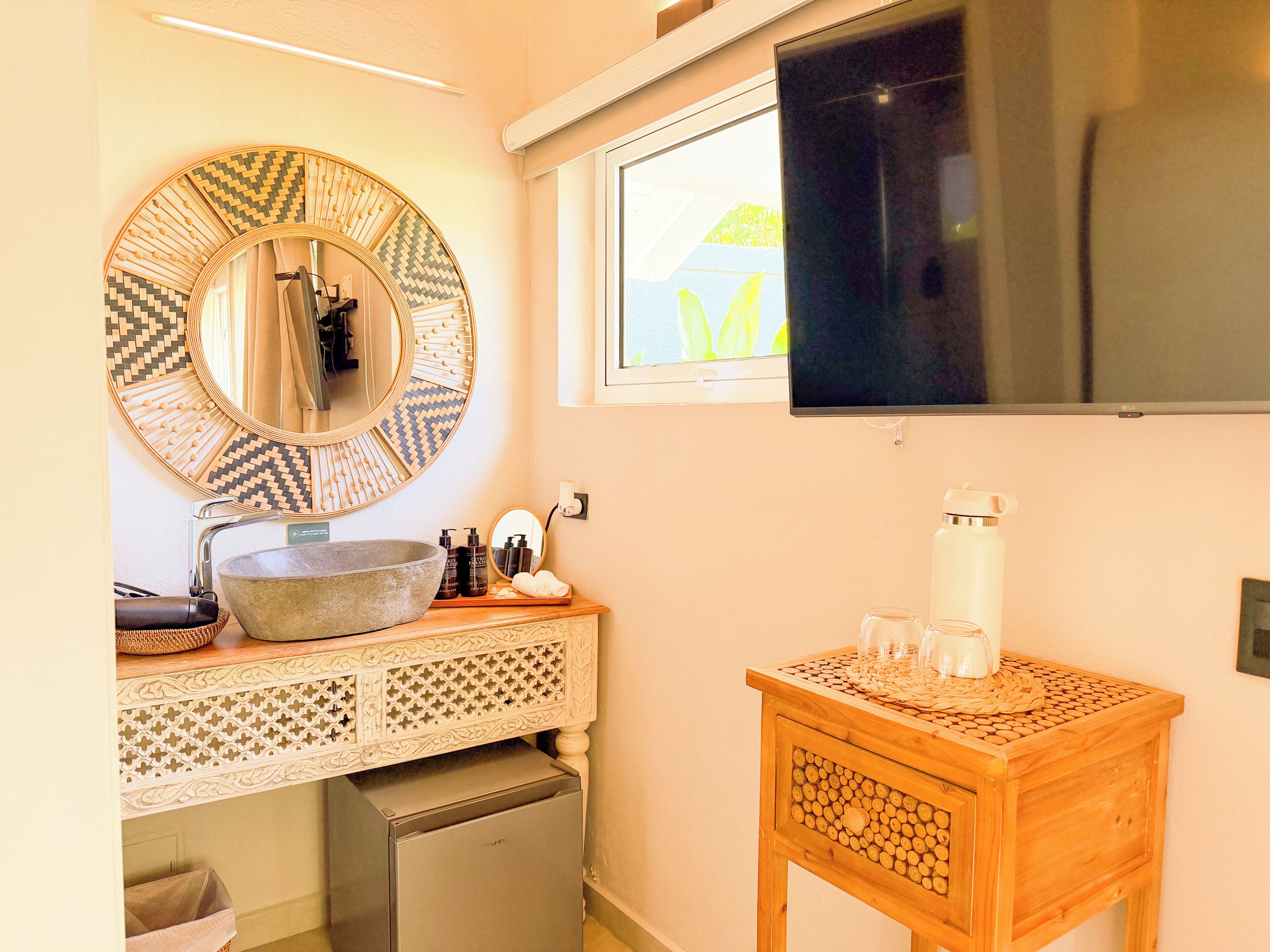 A bathroom with a round mirror, a sink, a small refrigerator, a wooden side table with a water bottle, and a window with a view.