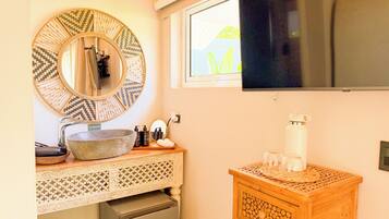 A bathroom with a round mirror, a sink, a small refrigerator, a wooden side table with a water bottle, and a window with a view.