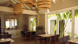 Un espace repas agrémenté de luminaires suspendus en osier, d’un grand miroir et offrant une vue sur des plantes tropicales.