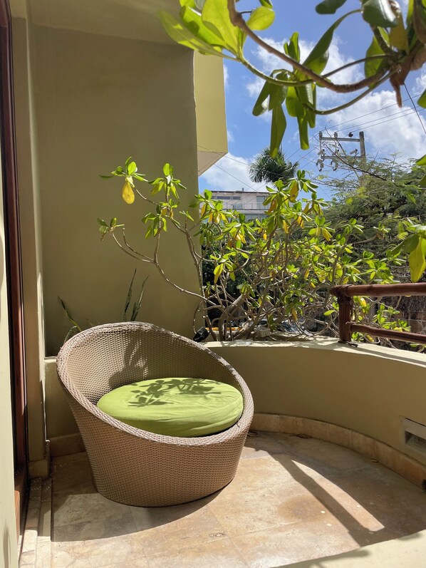 Una silla de mimbre con un cojín verde en un balcón con vistas a edificios y árboles circundantes.