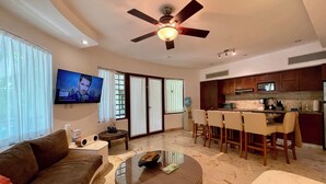 A modern living room with a sofa, a coffee table, a TV, and a kitchen area with a dining set. - 2BR Kobah • 1 Block to Beach & 5th - Pool+Gym (Playa del Carmen)