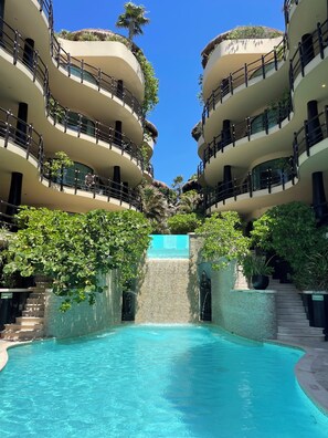 Un hotel de varios pisos con piscina central, balcones con plantas y una cascada.